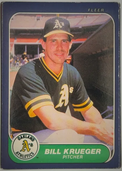 Front of 1986 Fleer Baseball Cards Bill Krueger #424, front of a baseball card. A dark blue/indigo border frames a color photograph of a player seated, wearing a dark Oakland Athletics cap with a white…
