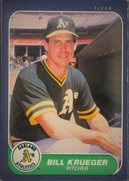 Front of 1986 Fleer Baseball Cards Bill Krueger #424, portrait baseball card front. A player in an Oakland Athletics uniform is shown seated in a stadium setting. The card has a dark navy-blue border, a…