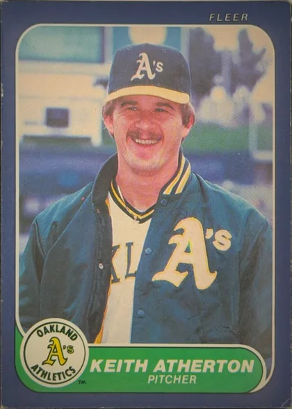 Front of 1986 Fleer Baseball Cards Keith Atherton #410, portrait baseball card front. A smiling Oakland Athletics player in a dark blue and yellow uniform occupies the central photograph. FLEER is printed…