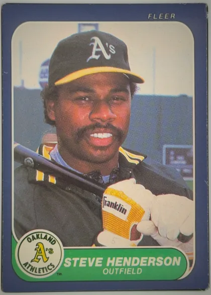 Front of 1986 Fleer Baseball Cards Steve Henderson #419 Base, front of a baseball card. A dark blue/indigo border frames a color photograph of a smiling Black male player wearing a black Oakland A's cap with a…