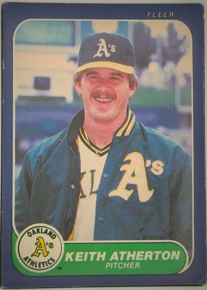 Front of 1986 Fleer Keith Atherton #410 Base, front of a baseball card. A dark blue border frames a color portrait of a smiling mustached man wearing a blue Oakland Athletics cap with a white…