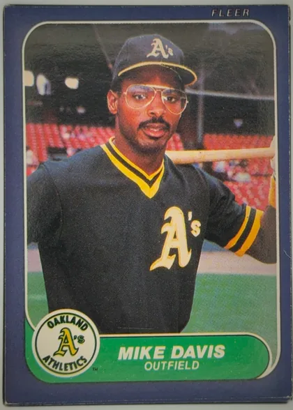 Front of 1986 Fleer Mike Davis #416 Base, portrait baseball card front. A player in an Oakland Athletics uniform and cap is shown in a close-up action portrait against a stadium background…