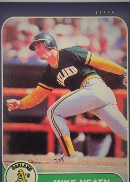 Front of 1986 Fleer Mike Heath #418 Base, front of a baseball card. A navy blue border surrounds a rounded-corner action photograph of a batter in an Oakland A's home uniform (black jersey…