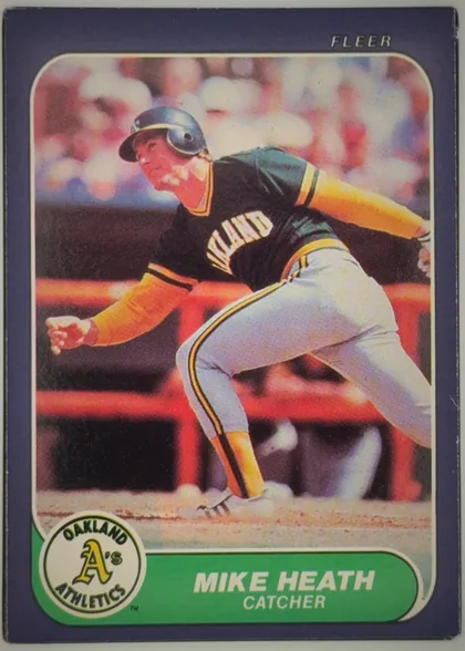 Front of 1986 Fleer Mike Heath #418 Base, front of a Fleer baseball card. A navy/dark-blue border with rounded inner corners surrounds a full-color action photograph of a batter in an…