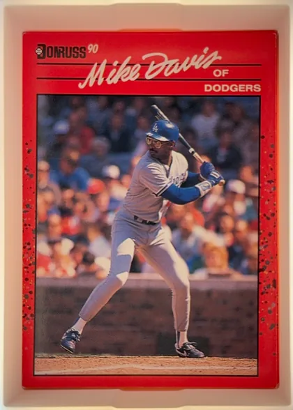 Front of 1990 Donruss Baseball Mike Davis #552, portrait-oriented baseball card front. A red speckled border surrounds a large action photograph of a batter in a Dodgers uniform. The upper left…