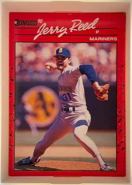 Front of 1990 Donruss Jerry Reed #614, portrait baseball card front. A red outer design surrounds a large action photograph of a Mariners pitcher in uniform delivering a pitch. The player…