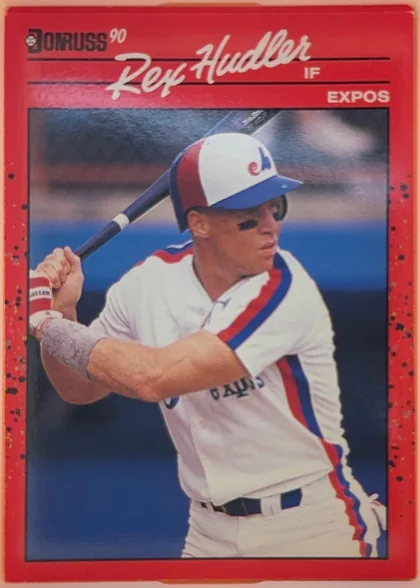 Front of 1990 Donruss Rex Hudler #366, portrait baseball card front. A red speckled border surrounds a large color action photograph of a batter in a white, red, and blue uniform. At the…