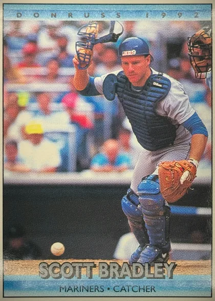 Front of 1992 Donruss Baseball Cards Scott Bradley #713, portrait baseball card front. A catcher in full protective gear is shown crouching near home plate with a baseball in the foreground and another…