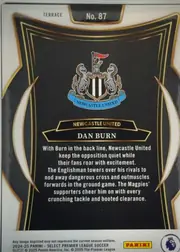 Back of 2024-25 Panini Select Premier League Soccer Dan Burn #87 Terrace, back of a portrait soccer card with a black and gold ornamental design. The Newcastle United crest is centered above a gold-edged nameplate reading…