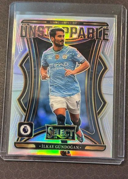 Front of 2024-25 Panini Select Premier League Soccer İLKAY GÜNDOĞAN #25 Prizm, front of a portrait soccer player card in a clear holder. A top banner reads 'MANCHESTER CITY' above a large blocky holographic title 'UNSTOPPABLE'…