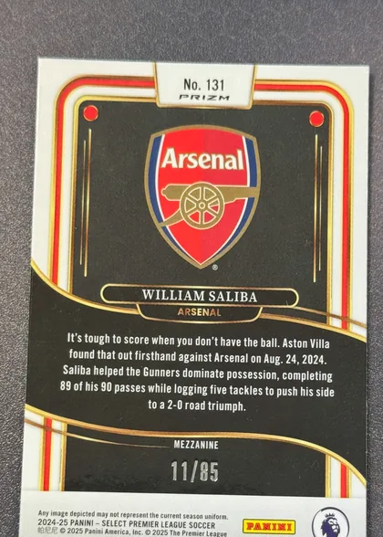 Back of 2024-25 Panini Select Premier League Soccer William Saliba #131 Mezzanine /85, serial numbered 11/85, portrait-oriented soccer card back with a black background and ornate cream, red, and gold curved border design. The Arsenal crest is centered in…