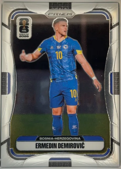 Front of 2026 Panini Prizm FIFA Ermedin Demirović #470 Base, portrait-format soccer card front. A full-body image of Ermedin Demirović in a blue Bosnia-Herzegovina kit occupies the center against a dark…