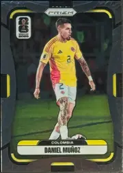 Front of 2026 Panini Prizm FIFA Soccer Cards Daniel Muñoz #180, portrait soccer card front. A player in a yellow Colombia uniform is shown full-length on a soccer field. The metallic PRIZM logo is centered near…