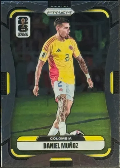 Front of 2026 Panini Prizm FIFA Soccer Cards Daniel Muñoz #180, portrait soccer card front. A player in a yellow Colombia uniform is shown full-length on a soccer field. The metallic PRIZM logo is centered near…