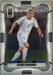 Front of 2026 Panini Prizm FIFA World Cup Soccer OTABEK SHUKUROV #451, portrait soccer card front. A full-body image of a player in a white Uzbekistan uniform occupies the center against a dark stadium background…