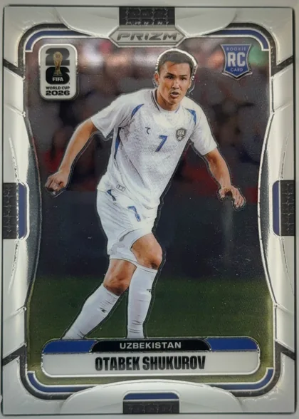 Front of 2026 Panini Prizm FIFA World Cup Soccer OTABEK SHUKUROV #451, portrait soccer card front. A full-body image of a player in a white Uzbekistan uniform occupies the center against a dark stadium background…