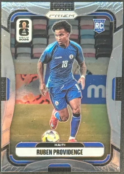 Front of 2026 Panini Prizm FIFA World Cup Soccer Ruben Providence #240 Rookie Card, upright portrait soccer card. A male soccer player in a blue Haiti kit, number 18, is shown running with a ball on a stadium field. The top contains…