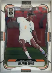 Front of 2026 Panini Prizm FIFA World Cup Soccer Wilfried Singo #256, portrait-format soccer card front. A large action photograph shows a player in a white Ivory Coast kit with orange number 5, kicking a ball on a…